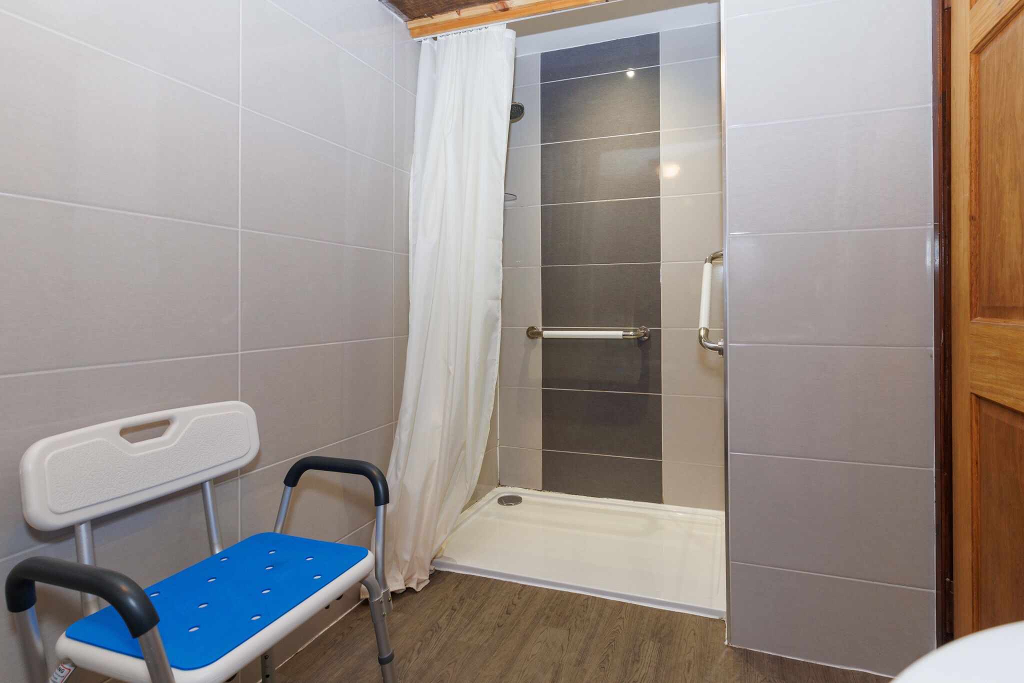 A shower area with a glass door, a white shower curtain, a grab bar, and a blue and white chair.