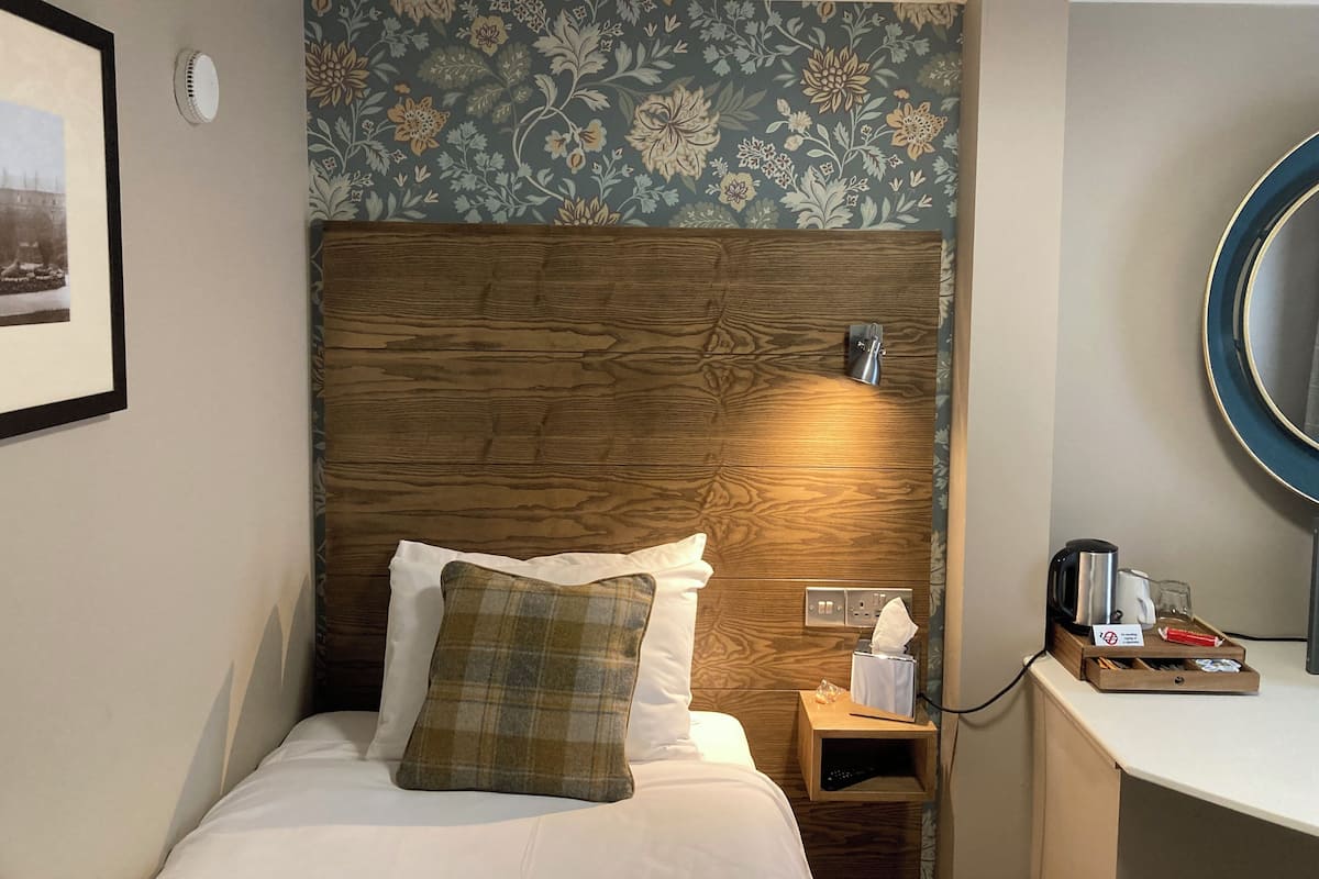 A hotel room with a wooden headboard, a bedside table with a lamp, a mirror, and a small tray with toiletries.