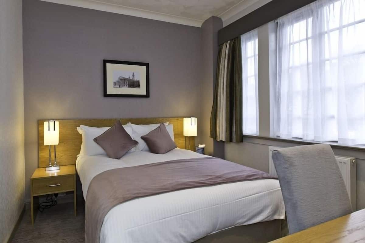 A hotel room with a bed, bedside lamps, a framed picture on the wall, a window with curtains, and a radiator.