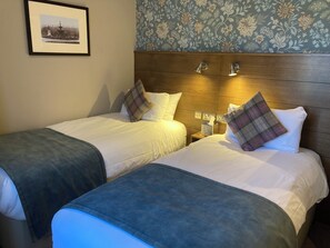 A hotel room with two beds, a wooden headboard, a nightstand with a lamp, and a framed picture on the wall.