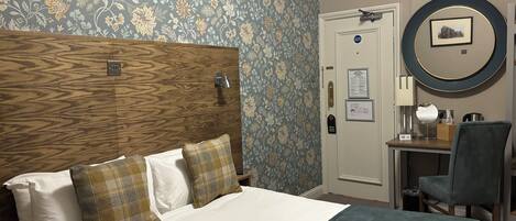 A hotel room with a wooden headboard, a bed with white and plaid bedding, a desk with a chair, a round mirror, and a door with a sign.