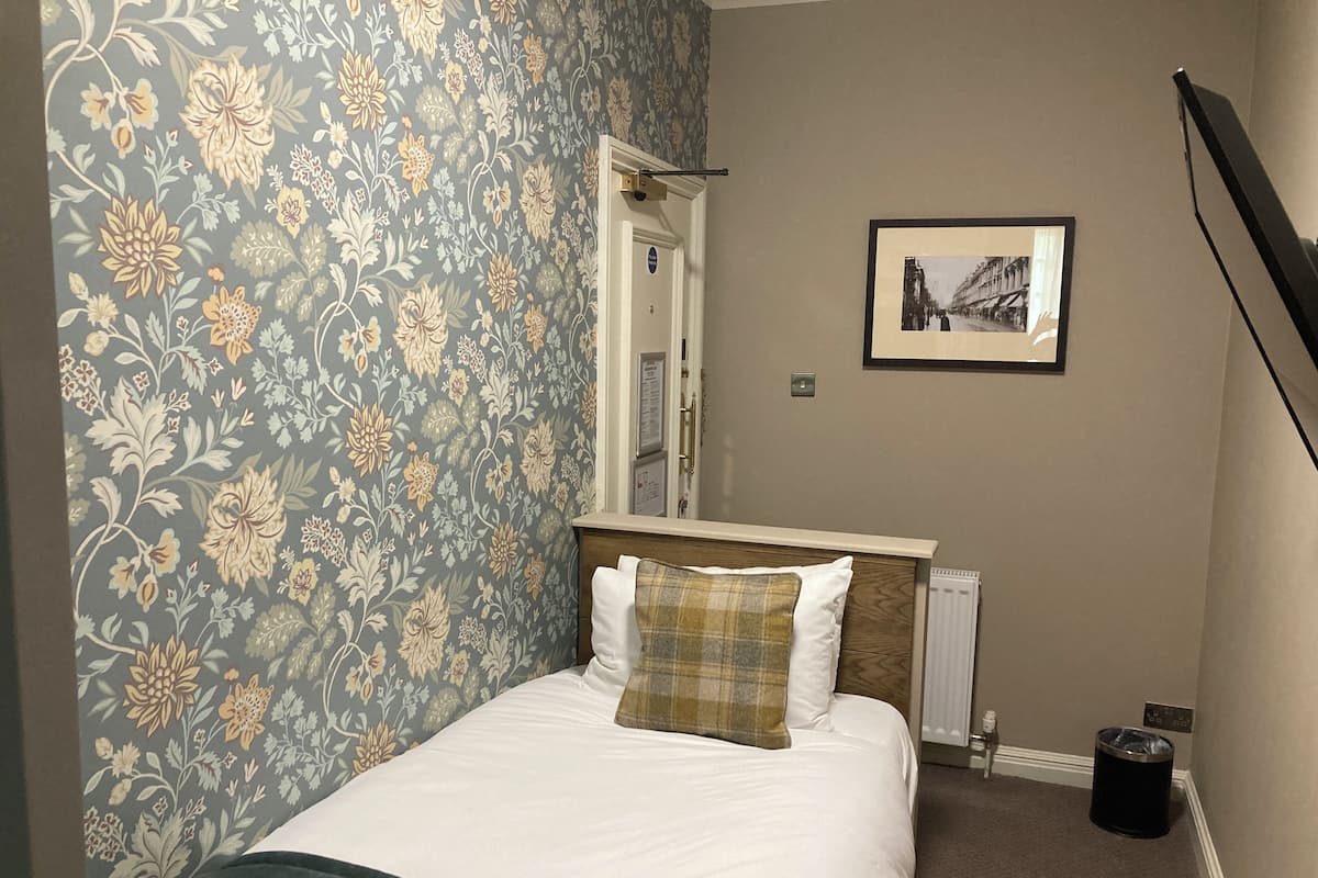 A neatly made bed with white and plaid bedding, a wooden headboard, and a floral wallpaper pattern on the left wall.