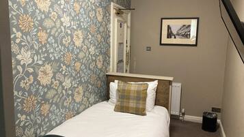 A neatly made bed with white and plaid bedding, a wooden headboard, and a floral wallpaper pattern on the left wall.