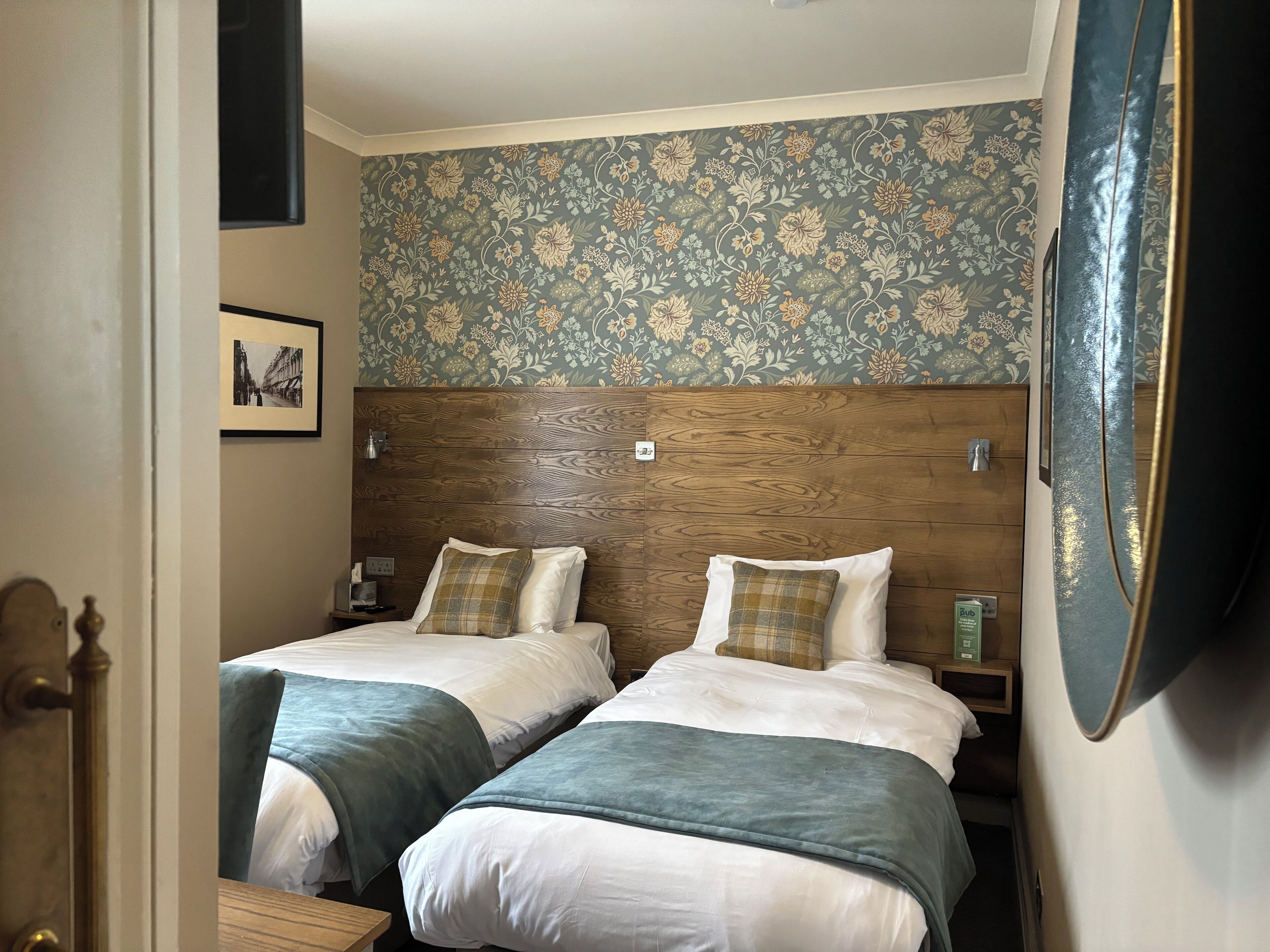 A hotel room with two beds, a wooden headboard, floral wallpaper, and a framed picture on the wall.