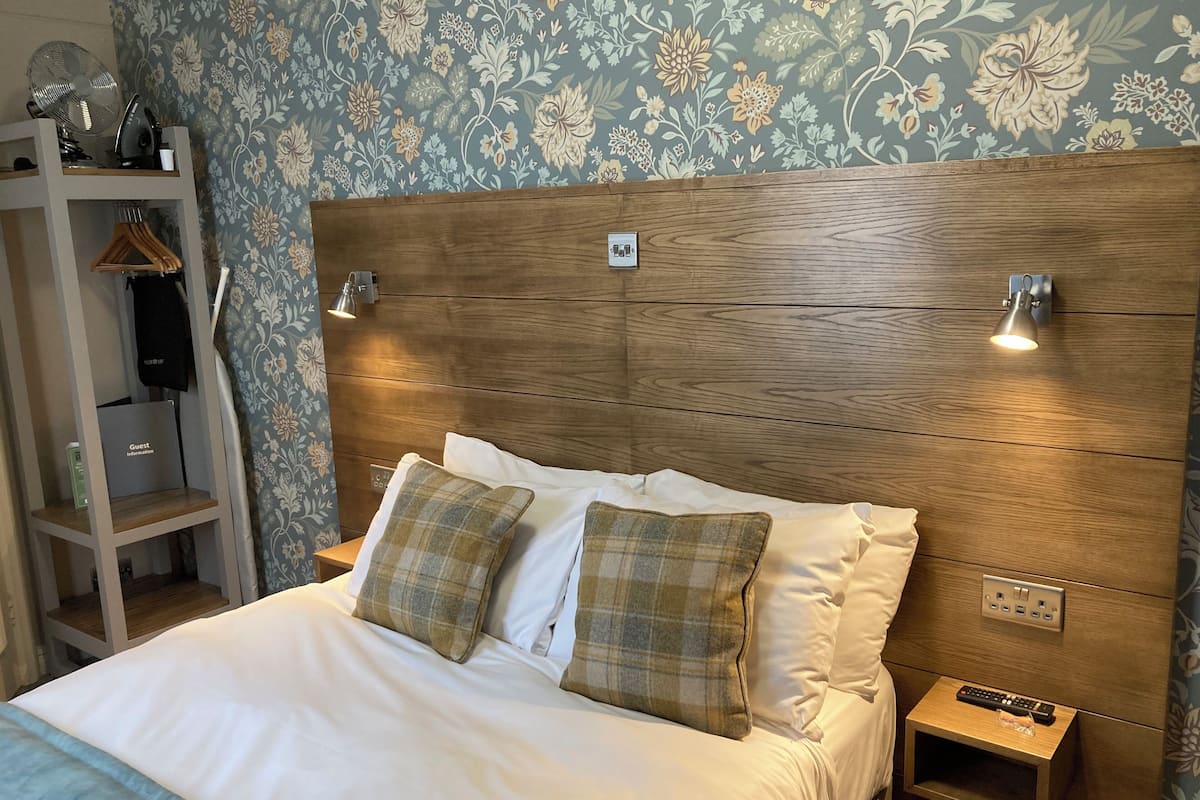 A neatly made bed with white linens and plaid pillows, a wooden headboard, bedside lamps, a small bedside table with a remote control, and a wall with floral wallpaper.