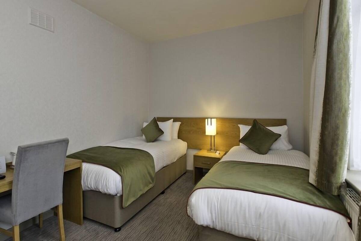 A hotel room with two single beds, a desk with a chair, a nightstand with a lamp, and a window with curtains.