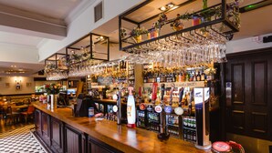 Zona de bar con mostrador de madera, estantes con botellas y una variedad de vasos colgando.