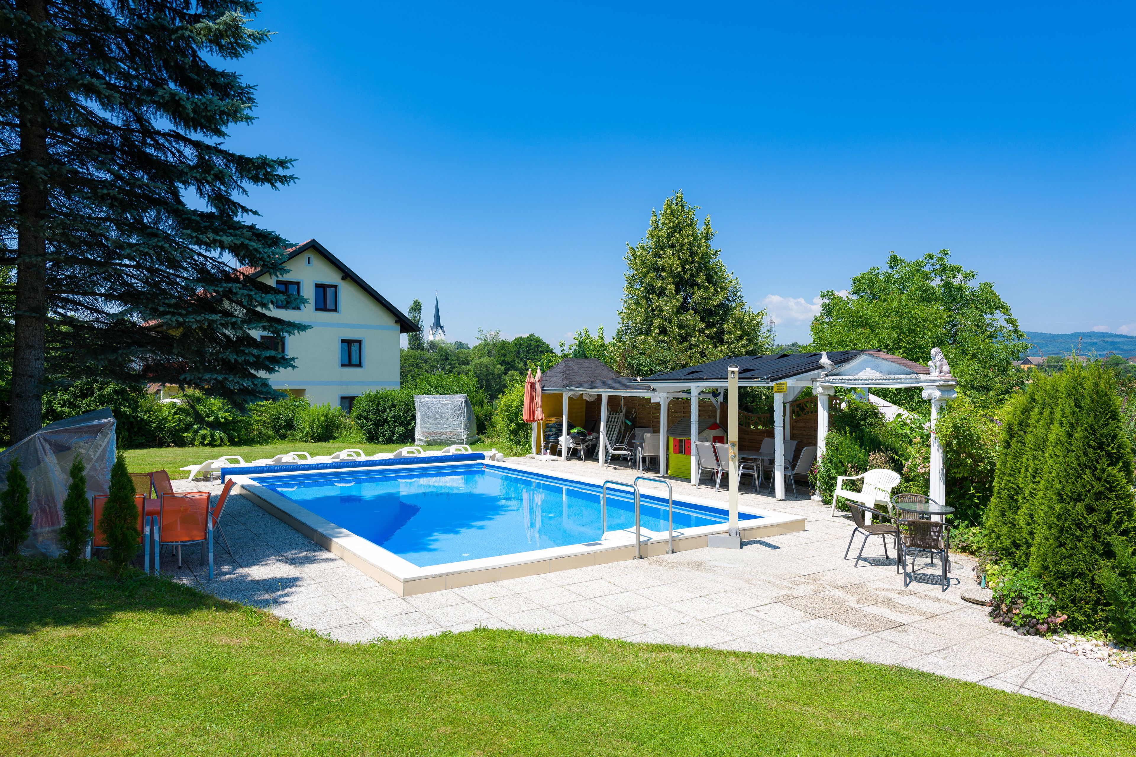 Una piscina con un porche techado, mobiliario exterior y una casa grande al fondo.