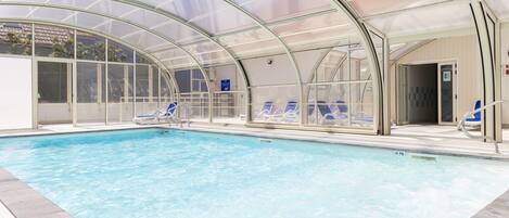 An indoor swimming pool with clear water, surrounded by blue lounge chairs and a glass-enclosed roof.