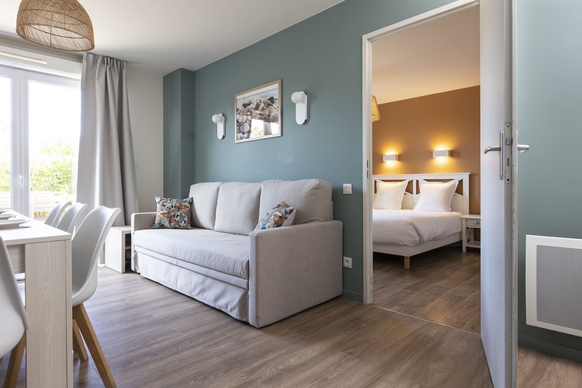 Ein modernes Wohnzimmer mit einer Couch, einem Esstisch und Stühlen. Im Hintergrund ist ein Bett mit Kopfteil und Nachttischlampen zu sehen.