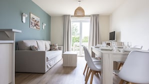 A modern living room with a sofa, dining table, and chairs. There is a television on a stand and a window with curtains. - Résidence Odalys Les Villas de la Baie (Le Crotoy)