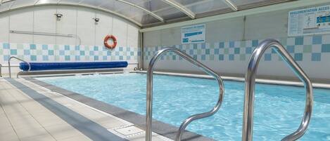 An indoor swimming pool with clear water, metal pool ladders, a blue checkered wall, and a glass ceiling.