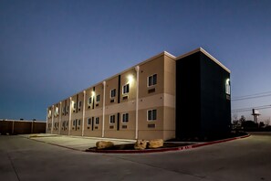 A multi-story hotel building with a flat facade, multiple windows, and a parking lot with a few cars and rocks. - Executive Inn Fort Worth West (Fort Worth)