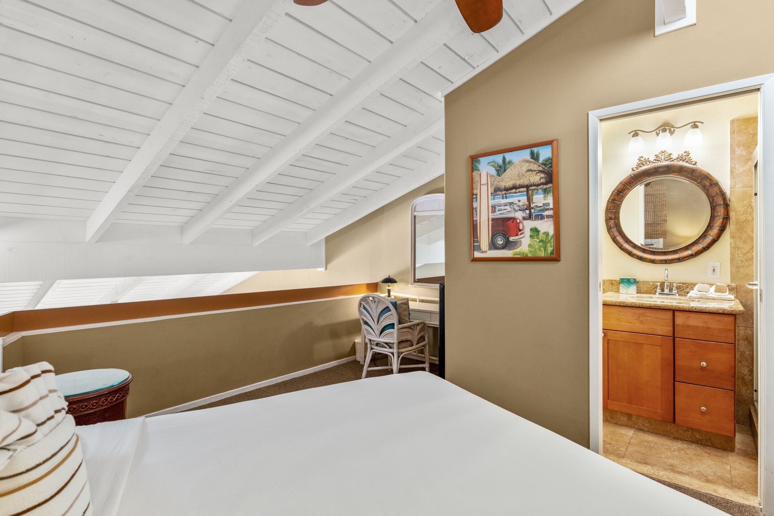 A bedroom with a bed, a desk, a chair, a bathroom with a mirror, and a painting on the wall.