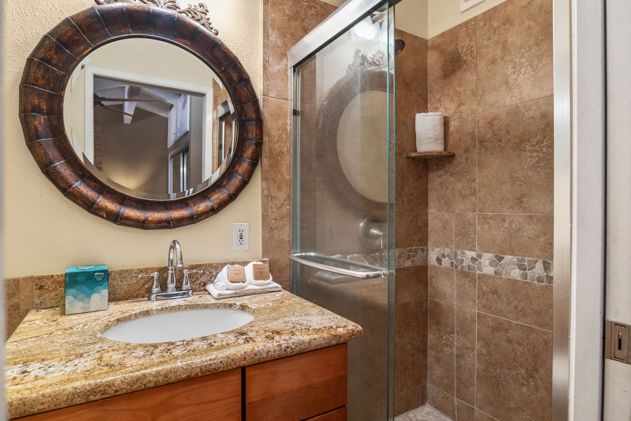 A bathroom with a granite countertop, a round mirror, a glass shower enclosure, and a tiled wall.