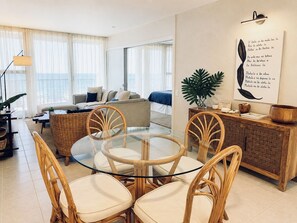 A modern living room with a glass dining table, wicker chairs, a sofa, and a large window with sheer curtains. - Kona Bali Kai 223 - Oceanfront 2 Bedroom Condo by RedAwning (Kailua-Kona)