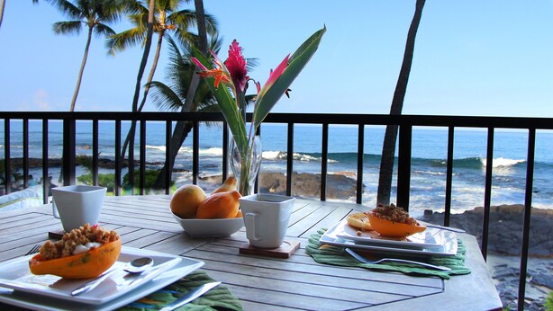 A table set with breakfast items, including a bowl of fruit, a cup, and a plate with food, overlooking a beach with palm trees and the ocean. - Kona Bali Kai 223 - Oceanfront 2 Bedroom Condo by RedAwning (Kailua-Kona)