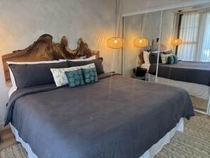 Una cama grande con colcha y almohadas de color oscuro, cabecera de madera y mesita de noche con lámpara.