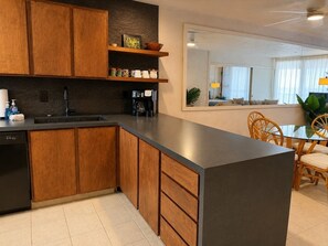 A modern kitchen with wooden cabinets, a black dishwasher, and a coffee maker. - Kona Bali Kai 223 - Oceanfront 2 Bedroom Condo by RedAwning (Kailua-Kona)