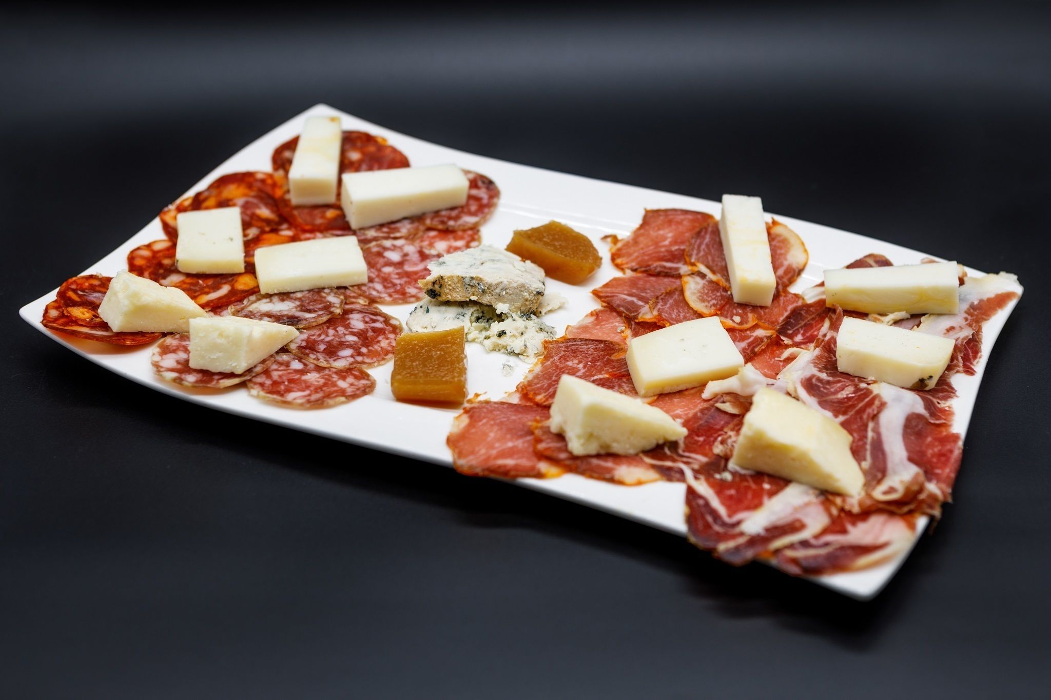A rectangular plate with various types of cured meats and cheese arranged on a dark background.