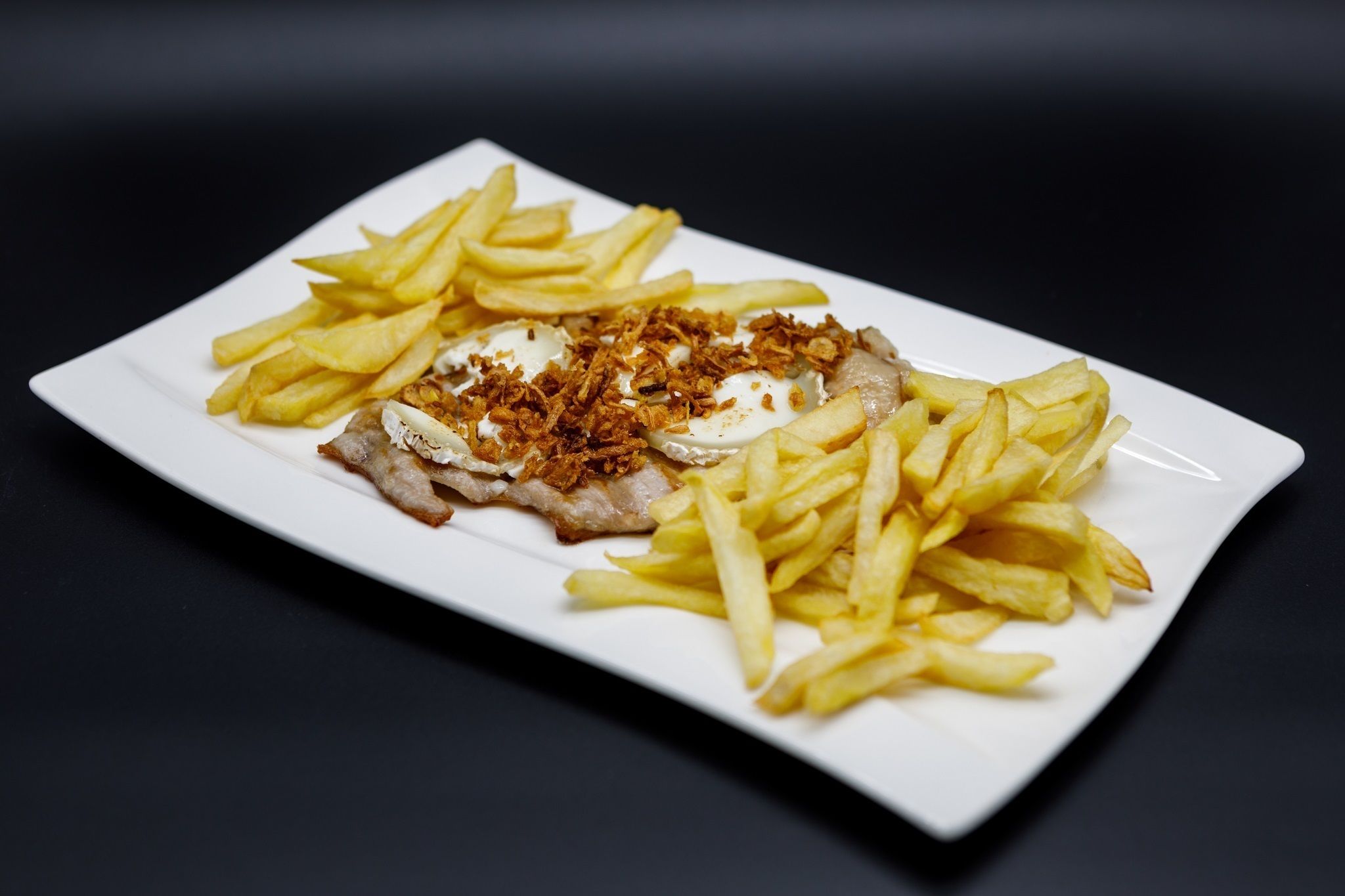 Um prato branco retangular com um pedaço de carne coberto com cebolas fritas e um pouco de molho, rodeado por batatas fritas.