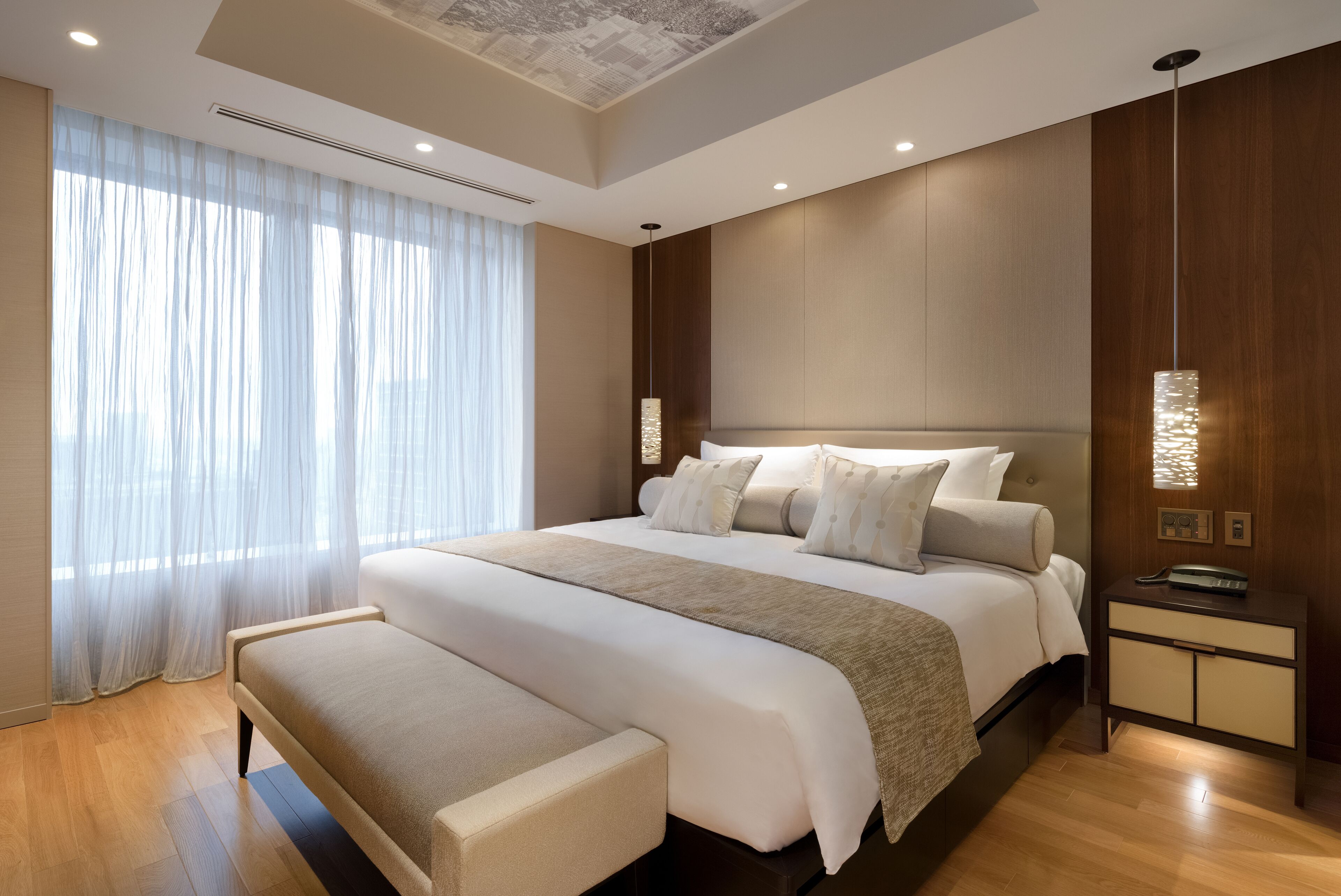A modern hotel room with a large bed, a bedside table, a bench, and a large window with sheer curtains.