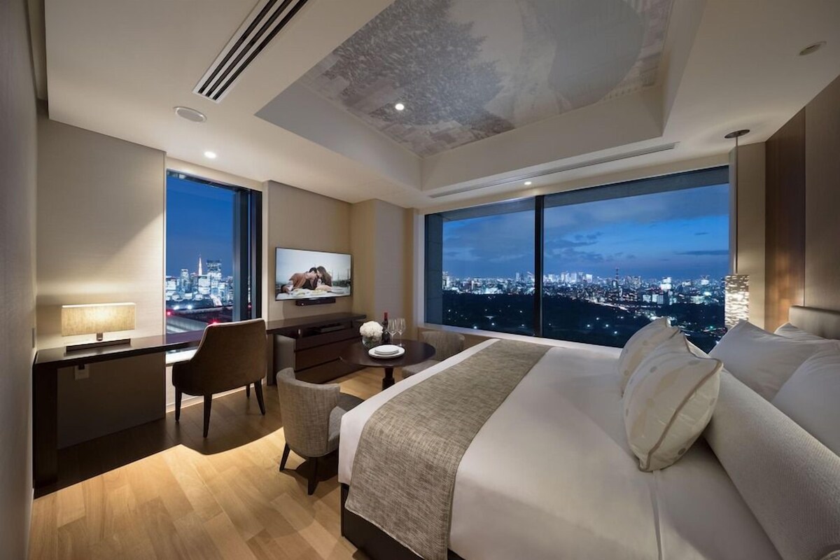 A modern hotel room with a large bed, a desk with a chair, a TV, and a city view.