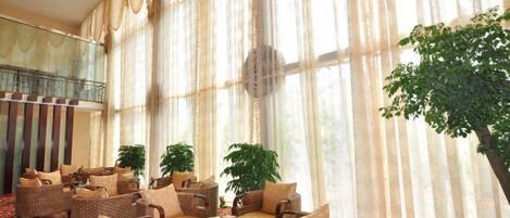 A spacious lobby with a high ceiling, large windows with sheer curtains, and a central seating area with wicker furniture.