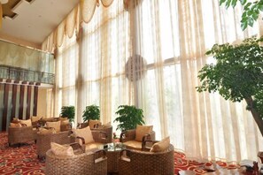 A spacious lobby with a high ceiling, large windows with sheer curtains, and a central seating area with wicker furniture.