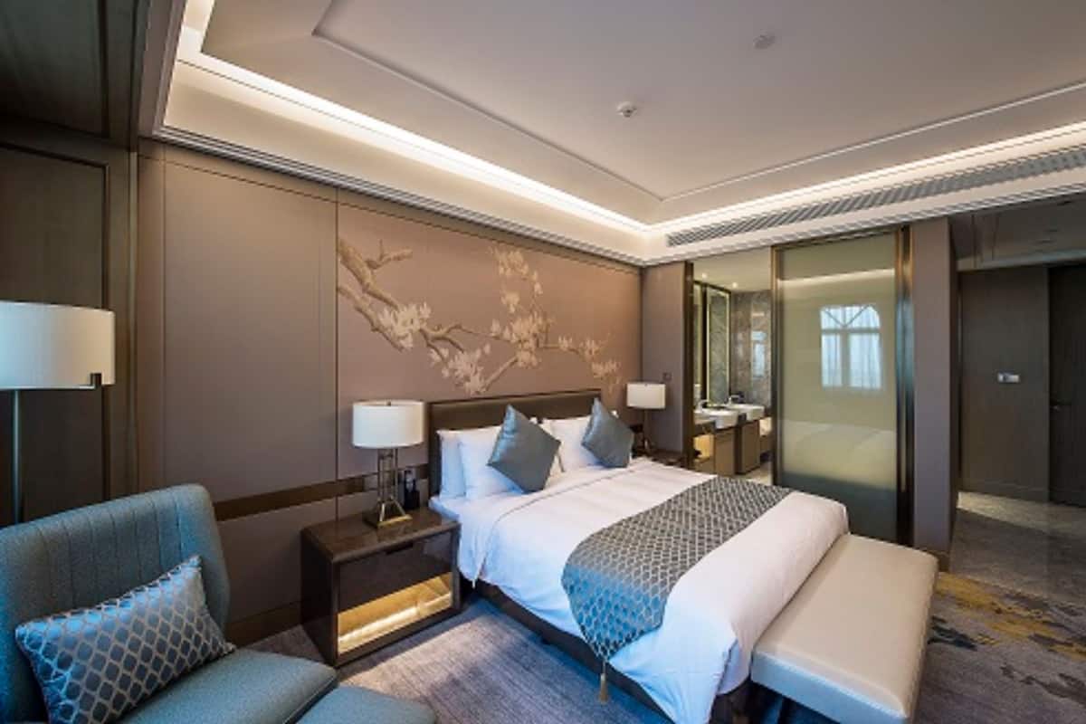 A modern hotel room with a large bed, a seating area with a sofa, a nightstand with a lamp, and a decorative wall piece.