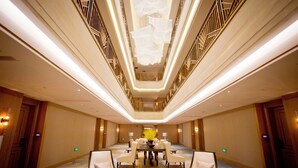 A spacious hotel lobby with a central dining area, a high ceiling with a geometric light fixture, and a patterned carpet.