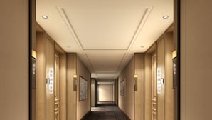 A long, narrow hotel hallway with a patterned carpet, wall-mounted light fixtures, and doors with room numbers.