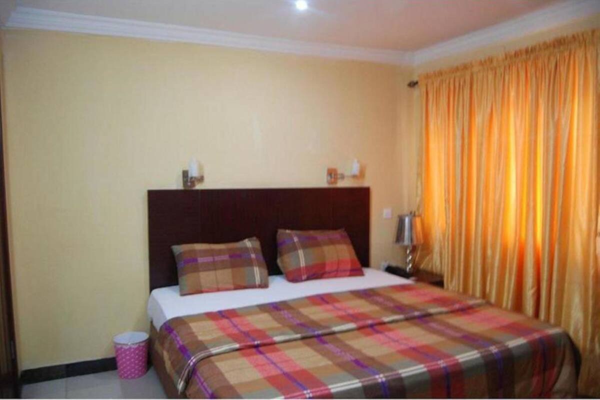 A hotel room with a bed, a headboard, a nightstand, a lamp, a curtain, and a trash bin.