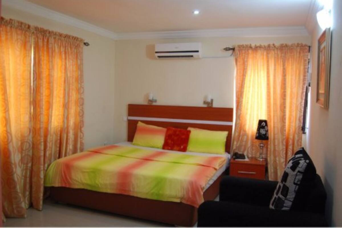 A bedroom with a bed, curtains, an air conditioner, a bedside table, and a lamp.