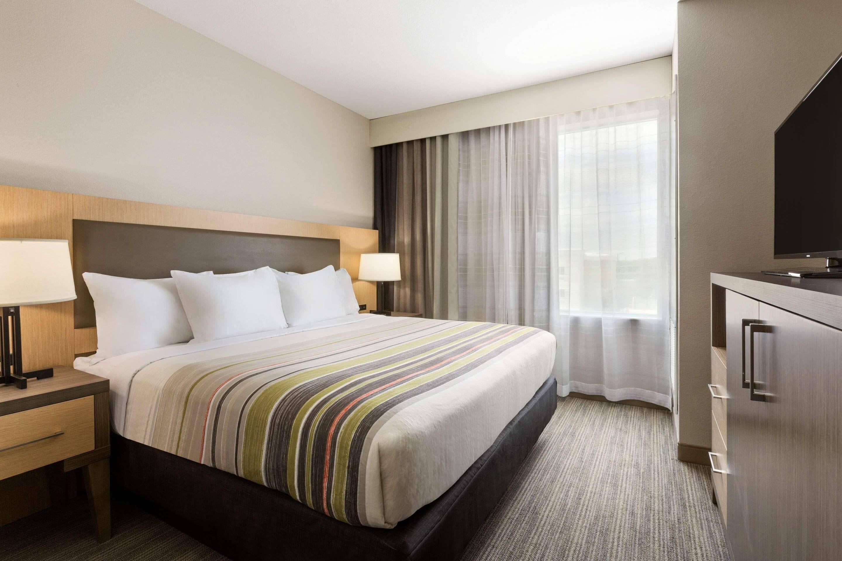 A hotel room with a large bed, bedside lamps, a flat-screen TV, a desk, and a view of the outdoors through curtains.