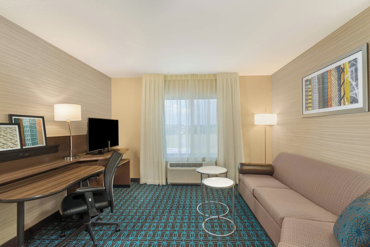 A hotel room with a desk, chair, sofa, two side tables, a TV, and a window with curtains.