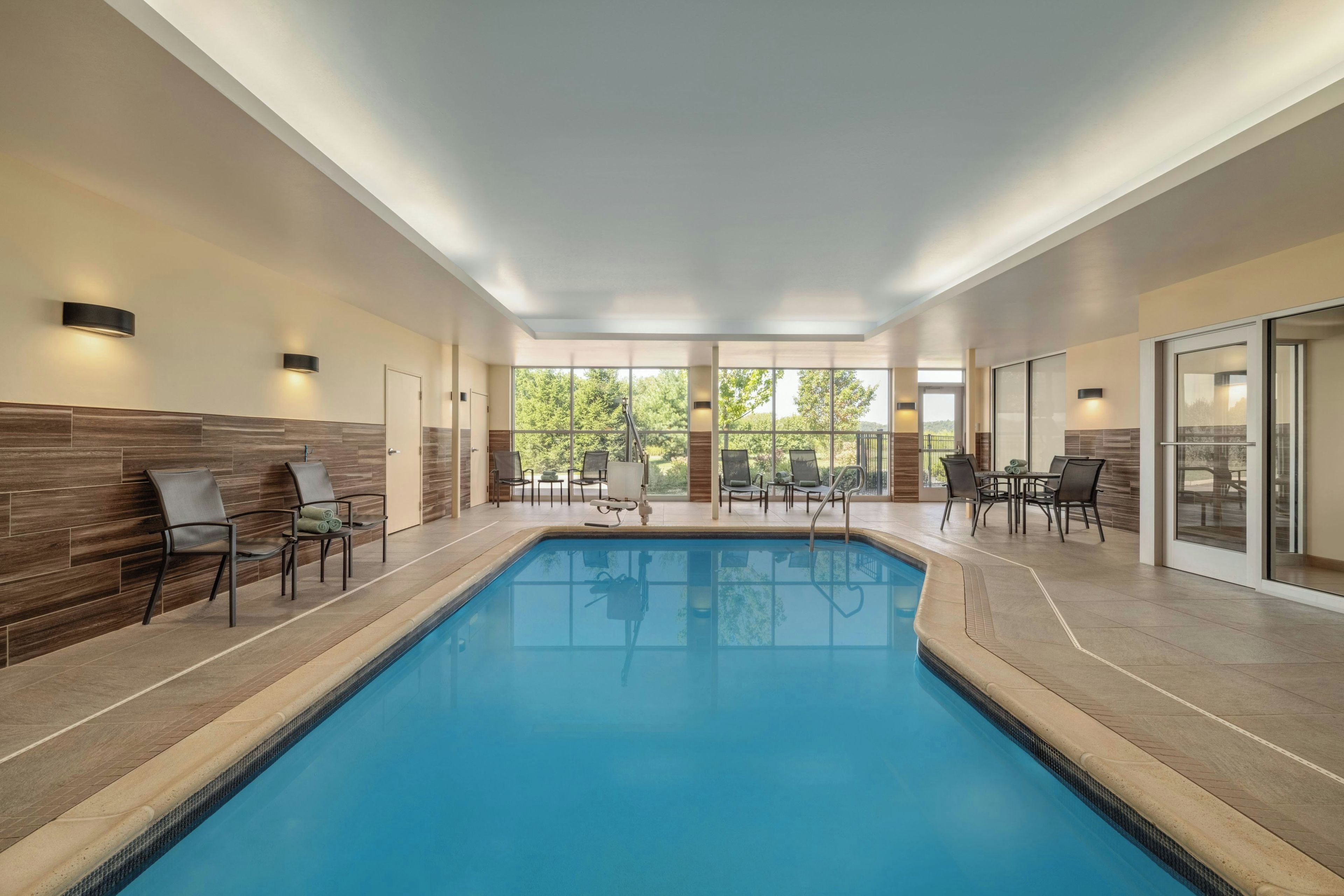 An indoor swimming pool with clear blue water, surrounded by lounge chairs and tables, with large windows offering a view of greenery outside.