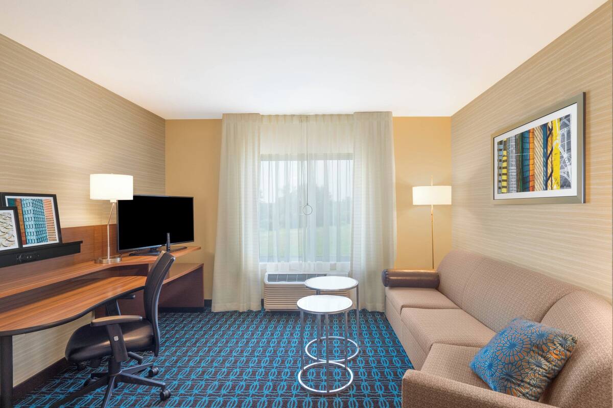 A hotel room with a desk, a chair, a sofa, a TV, a lamp, a window with curtains, and a framed picture on the wall.