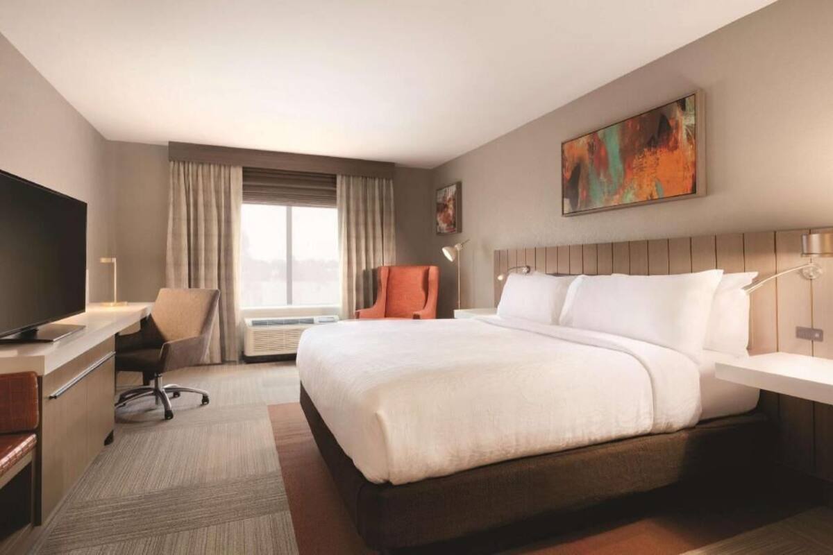 A hotel room with a bed, desk, chair, TV, and artwork on the wall.