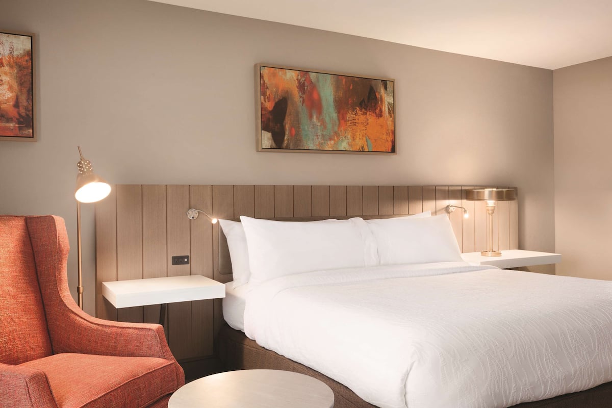A hotel room with a bed, a nightstand, a lamp, a chair, and a painting on the wall.