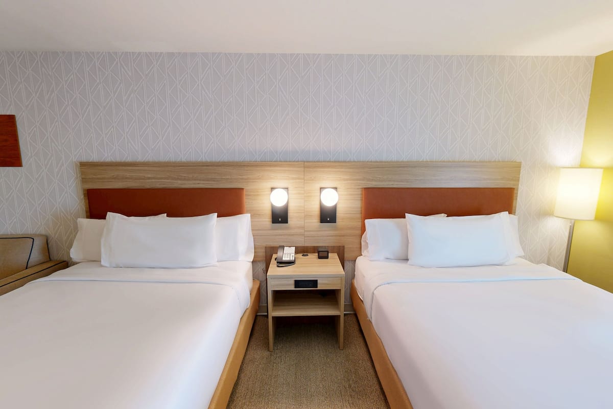 A hotel room with two beds, a wooden headboard, bedside lamps, a nightstand, and a wall-mounted artwork.