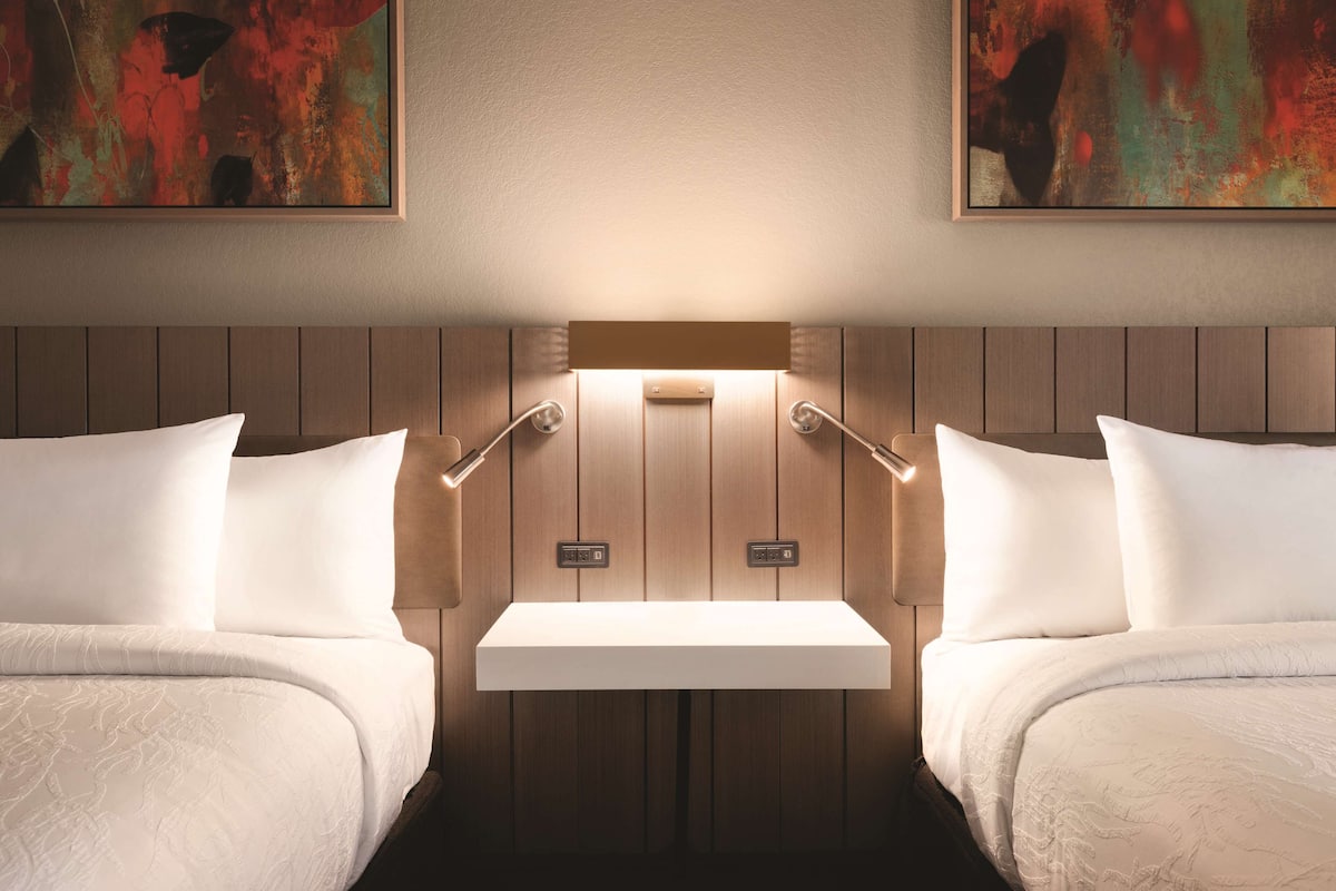 A hotel room with two beds, a bedside table, and a wall-mounted shelf.