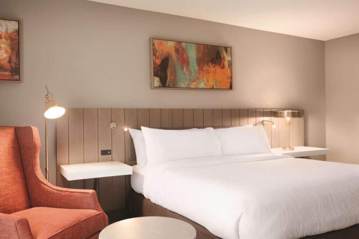 A hotel room with a bed, a chair, a bedside table, a lamp, and a painting on the wall.