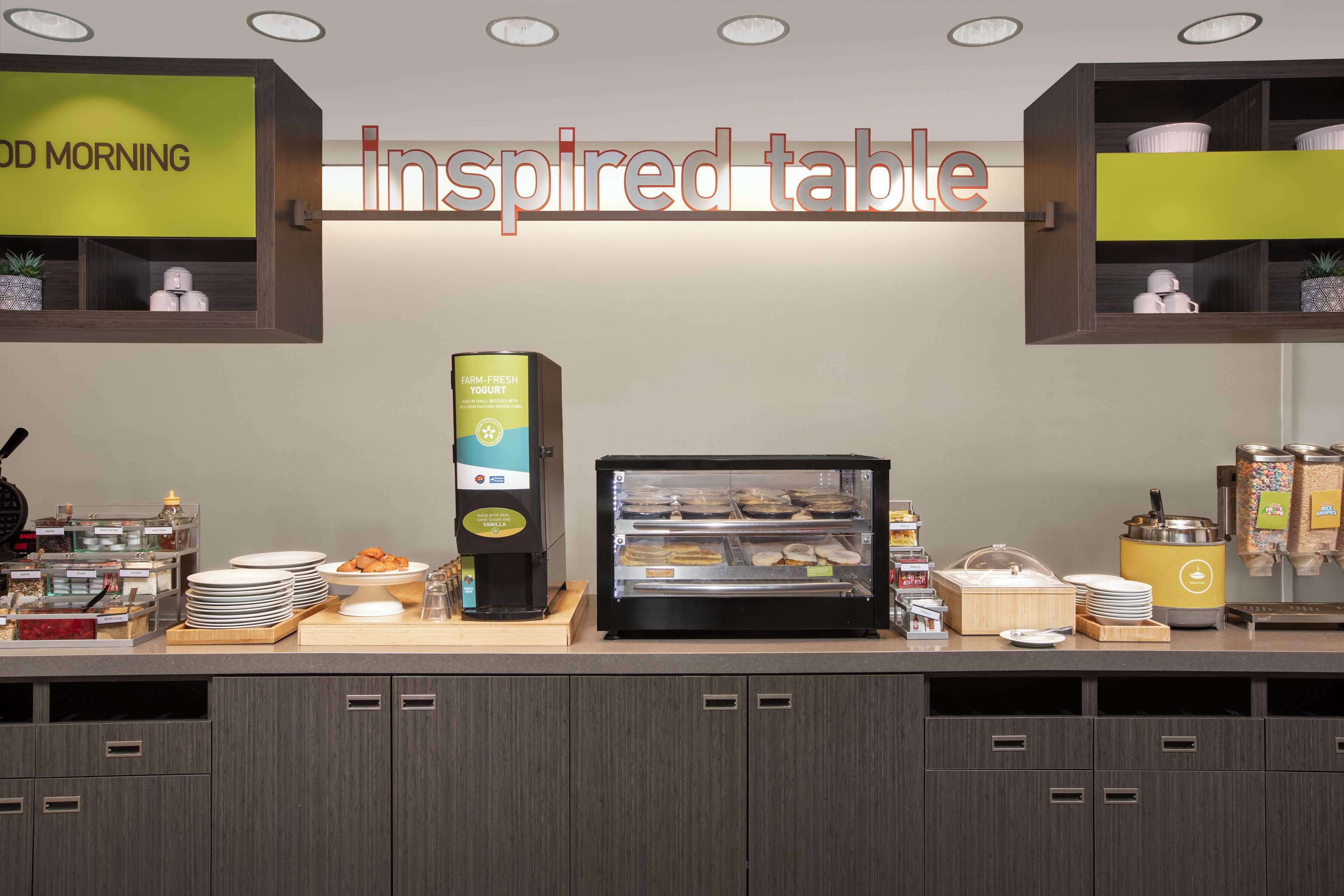 Espaço de buffet de café da manhã com uma placa indicando "inspired table", uma vitrine com doces e uma máquina de café.
