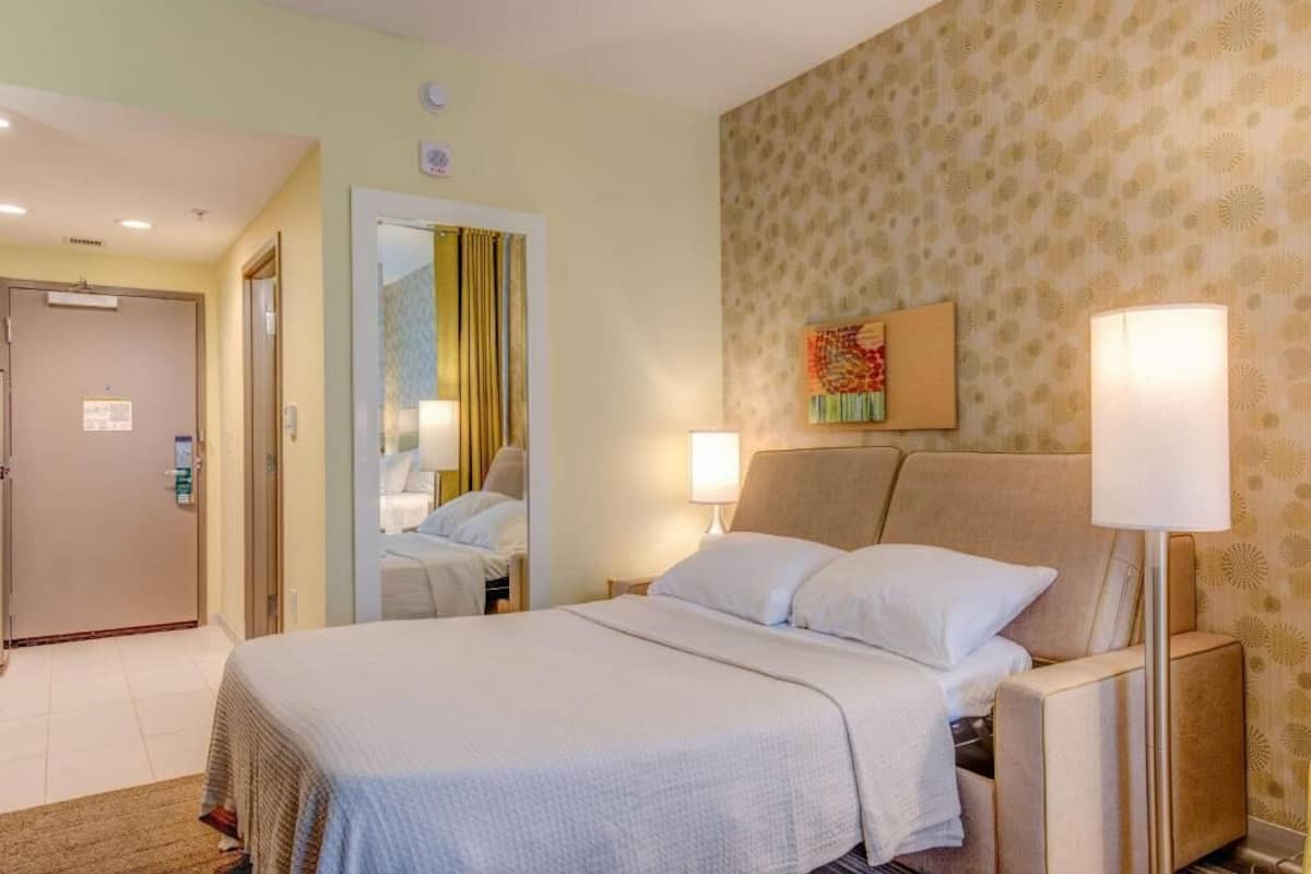 Habitación de hotel con cama, lámparas de noche, un espejo, una puerta y una pared con papel tapiz estampado.