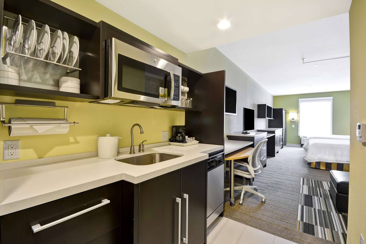 Una cocina moderna con microondas, pileta y muebles. Hay un pequeño comedor con mesa y sillas. La habitación tiene un ventanal grande y una cama al fondo.