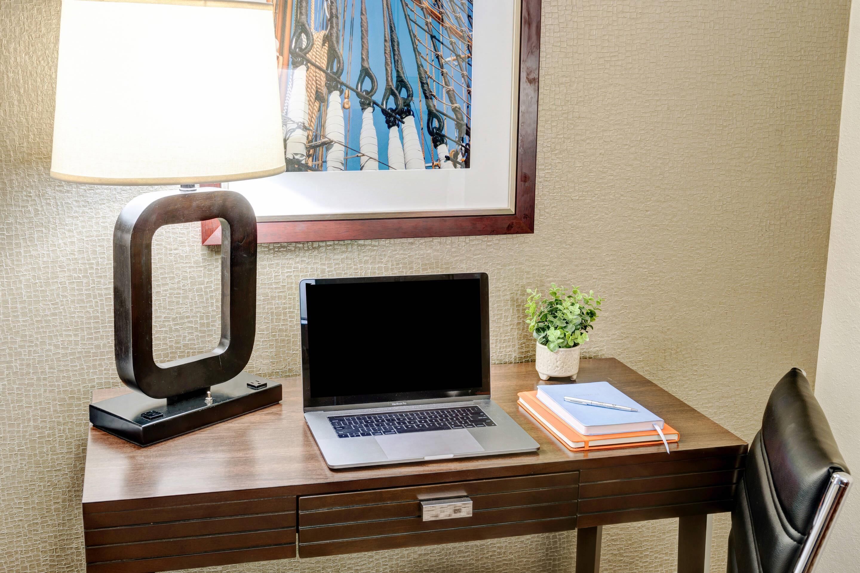 A wooden desk with a laptop, a potted plant, and a framed artwork.