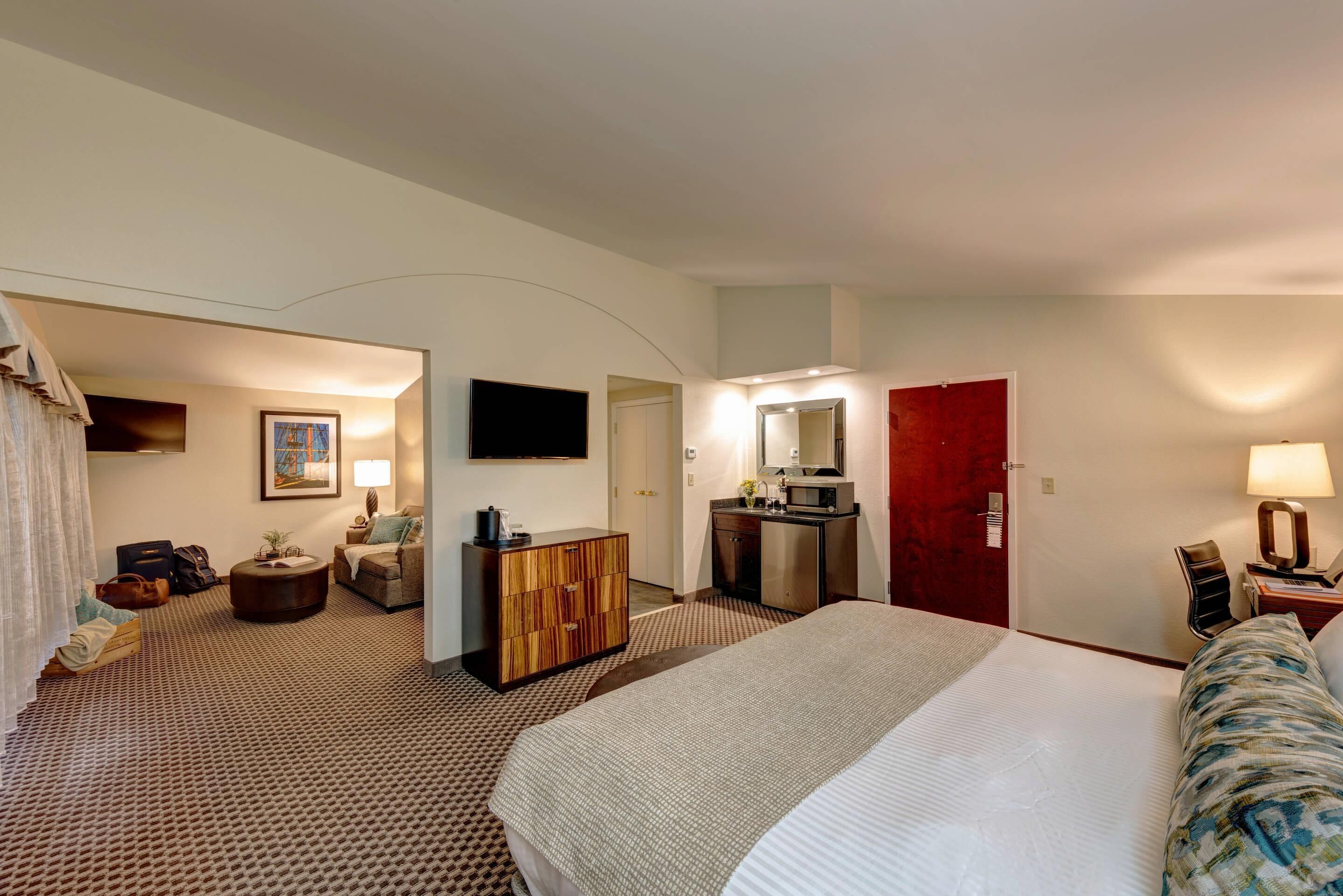 A hotel room with a large bed, a desk with a computer, a TV, and a red door.