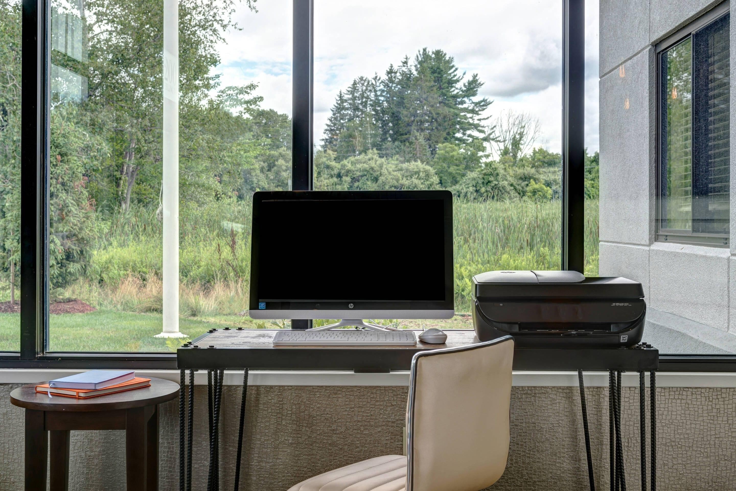 A modern office with a desktop computer, printer, and chair, overlooking a lush green landscape through large windows.
