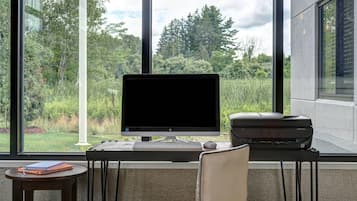 A modern office with a desktop computer, printer, and chair, overlooking a lush green landscape through large windows.